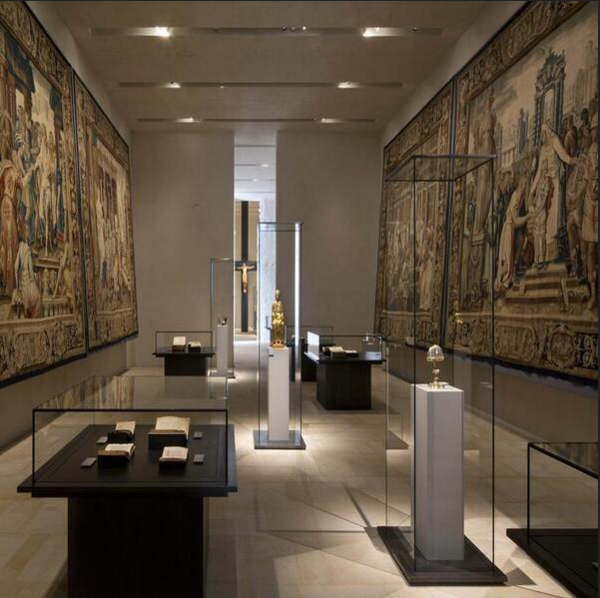 Ein stimmungsvoll beleuchteter Ausstellungsraum des Dommuseums. Unter hohen, schlanken Glasvitrinen werden kleine Objekte auf wei&szlig;en Sockeln ausgestellt. An den W&auml;nden rechts und links sieht man Wandgem&auml;lde oder Wandteppiche.