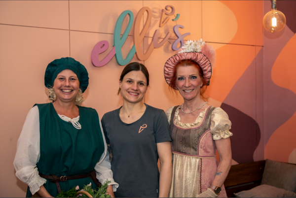 Drei Frauen stehen unter dem bunten Schriftzug "Elli's". Die Frau in der Mitte ist die Eisverkäuferin und rechts und links stehen die verkleideten Kostümführerinnen.