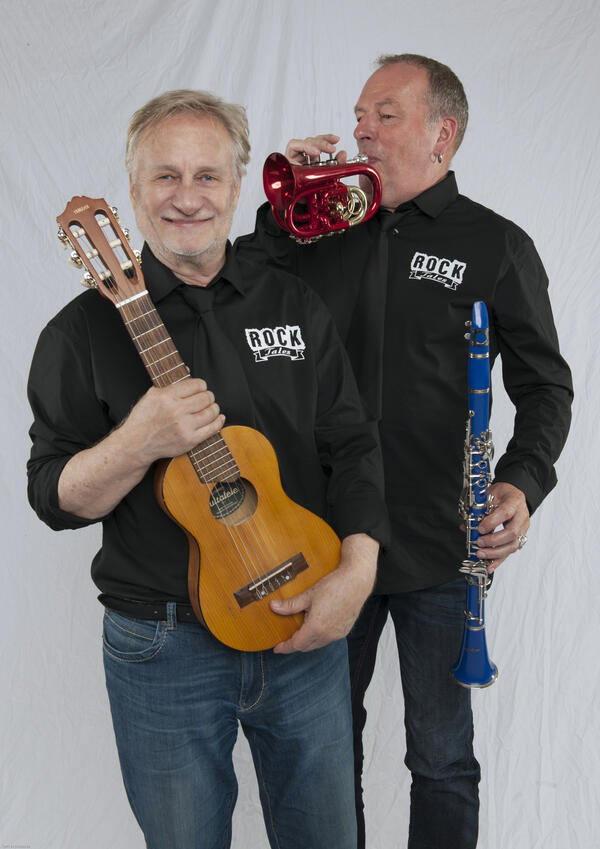 Zwei weißhaarige Männer, einer mit einer kleinen Akustikgitarre, der andere mit einer kleinen Trompete und einer Klarinette in der Hand.