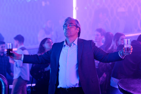 Ein Mann in Anzug und mit Brille steht, die Arme ausgebreitet, in einem mit lila Neonlicht beleuchteten Raum und hat die Augen geschlossen