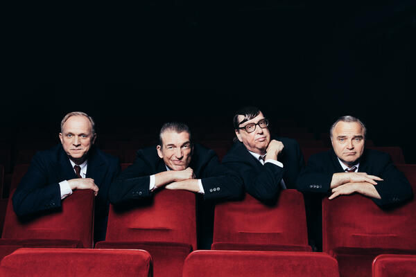 Vier weiße Männer in schwarzen Anzügen lehnen über roten Stuhllehnen vor einem schwarzen Hintergrund. Ähnlich wie in einem Kino oder Konzertsaal.