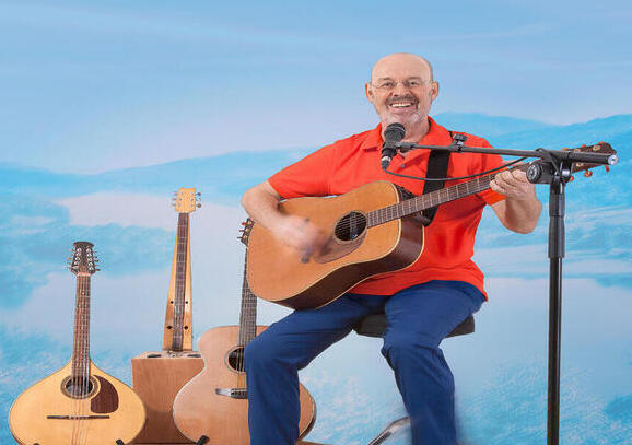 Ein Mann mit orangenem Oberteil und Jeans sitzt auf einem hohen Hocker vor einer Wand mit Wolken, eine Akustik-Gitarre in der Hand und andere Akustikinstrumente im Hintergrund. 