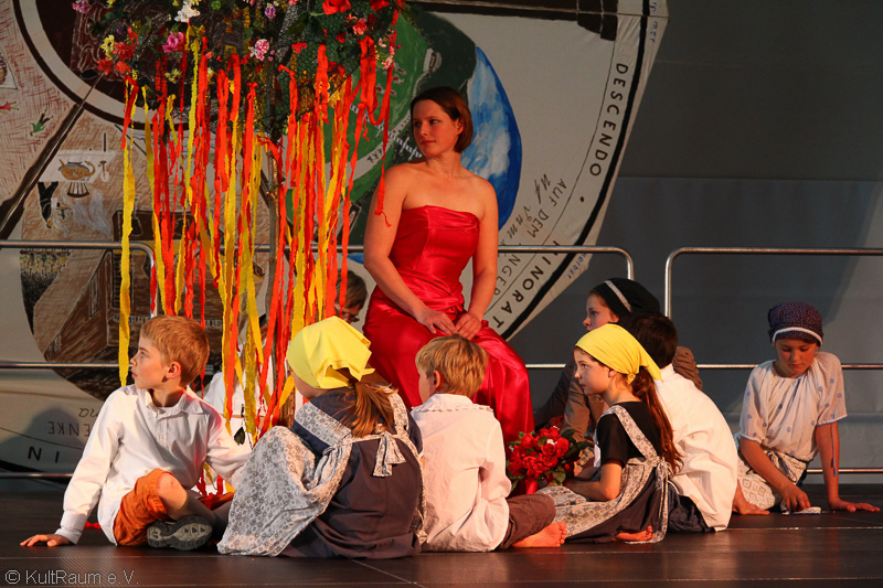 Bild vergrößern: Eine Bühne auf der eine Frau im roten Kleid und Kinder in Kostüm sitzen.