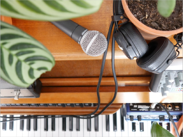 Ein Mikrophon und Köpfhörer liegen auf einem Klavier. Ein Stück einer Pflanze ist auch zu sehen. Das Bild wirkt hell und einladend.